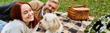 A man and woman relax on a cozy blanket with their dog in a park. clipart