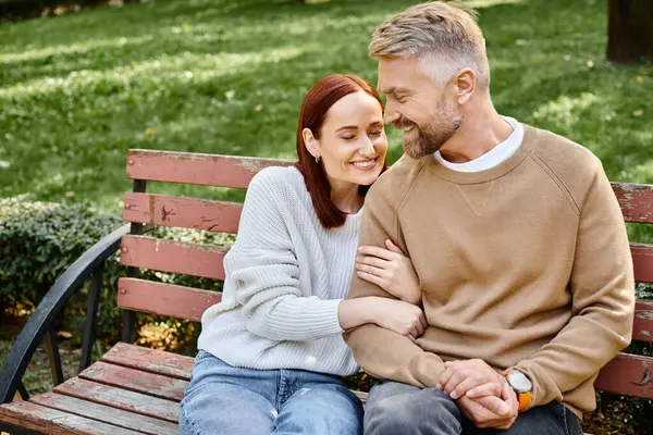 stock image A man and a woman in casual attire sit together on a wooden bench in a park.