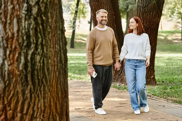 stock image A man and woman in casual attire walk together in a peaceful park setting.