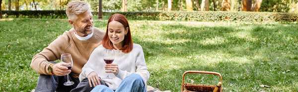 Adult man and woman sitting on a blanket, holding wine glasses in a park.