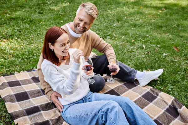 A man and woman sit together on a blanket, holding wine glasses.
