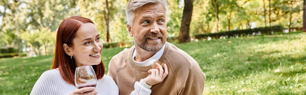 A man and a woman in casual attire, holding wine glasses, enjoying a romantic stroll in the park.