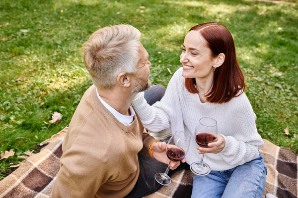 A man and a woman sit on a blanket, holding wine glasses in a park.