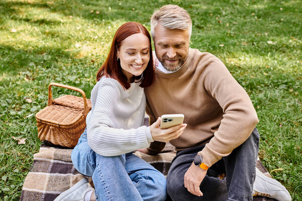 Пара, сидящая на одеяле, поглощенная мобильным телефоном.