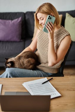 Kısa saçlı şık bir kadın koltukta dinleniyor, cep telefonuna odaklanmış haldeyken tatmin olmuş kedisi kucağında dinleniyor..
