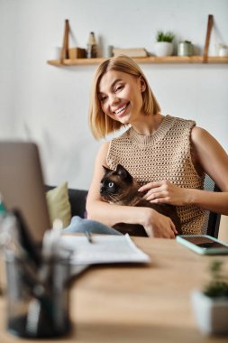 Masasında oturan kısa saçlı rahat bir kadın, kucağında bir kedi, evinde huzurlu bir anı paylaşıyor..