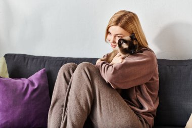 Kısa saçlı bir kadın bir kanepede dinlenir, birlikte sakin bir anı paylaşırken kedisini kucağına alır..