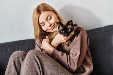 Kısa saçlı bir kadın kanepede uzanıyor, sevgili kedisini şefkatle tutuyor..