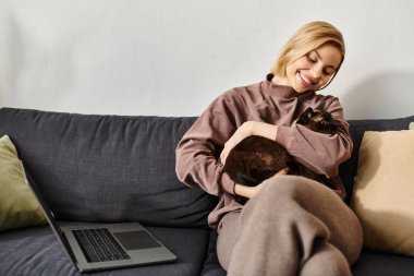 Kısa saçlı bir kadın kanepede rahatlar, kedisini kollarına alır. Sessiz bir yakınlık ve rahatlık anlarını paylaşıyorlar..