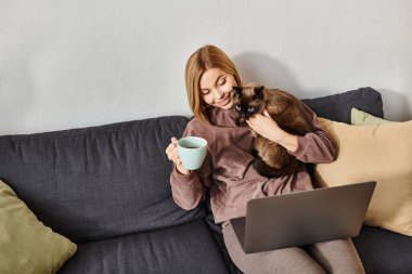 Kısa saçlı bir kadın bir kanepede dinleniyor, bir fincan kahvenin tadını çıkarırken bir kediyi kollarında tutuyor..