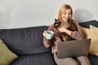 Kısa saçlı bir kadın kanepede oturmuş, kedisini kollarında tutarken bir fincan kahvenin tadını çıkarıyor..
