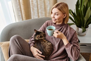 Kısa saçlı bir kadın kanepede oturuyor, kucağında kedi beslerken bir fincan kahve içiyor..