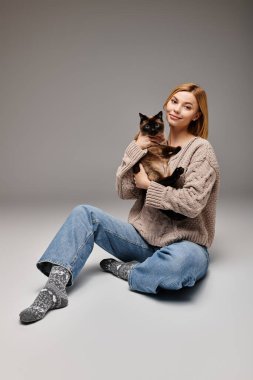Kısa saçlı bir kadın yerde oturuyor, sevgiyle kucağında bir kedi tutuyor..