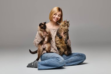 Kısa saçlı bir kadın yerde oturuyor, iki kediyi kollarına alıyor, evde uyumlu bir bağ kuruyor..