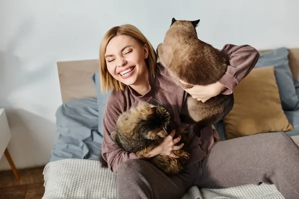 ベッドの上に座っている短い髪の女性は 優しく彼女の腕に2匹の猫を抱き 自宅で平和な瞬間を楽しんでいます — ストック写真