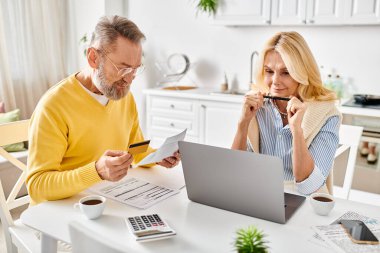 A mature couple in cozy homewear deeply engrossed in reviewing paperwork together at a table in their kitchen. clipart