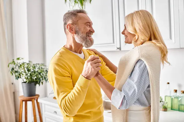 stock image A mature loving couple in cozy homewear dancing joyfully together in a warm kitchen setting.
