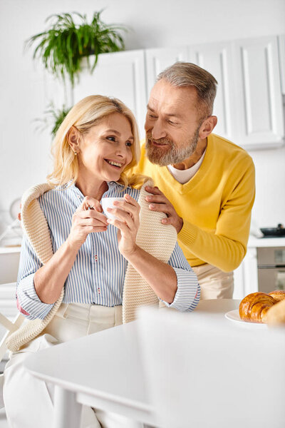 A mature man and woman in cozy homewear sit together at a kitchen counter, enjoying a peaceful moment together.
