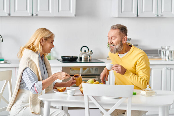 A mature loving couple in cozy homewear enjoying a meal together at a table in their kitchen at home.