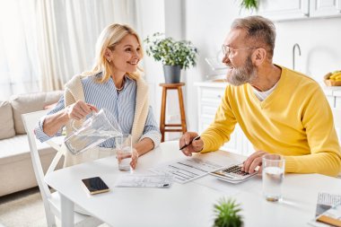 Masada oturup hesap makinesini finansal hesaplamalar için kullanmaya odaklanmış rahat bir ev elbisesi içinde olgun bir erkek ve kadın..