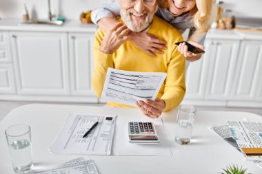 A mature man and woman in cozy homewear sit at a table working on a calculator, seemingly budgeting and planning together. clipart