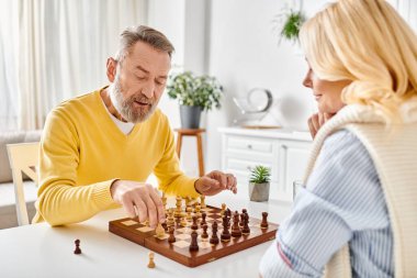 A mature, loving couple in cozy homewear engaged in a competitive game of chess, focused on the board before them. clipart