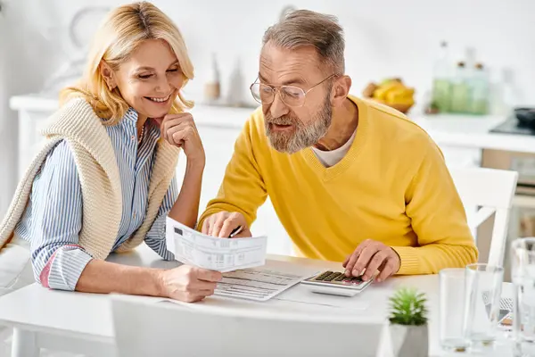 stock image A mature man and woman in cozy homewear are seated in the kitchen, engrossed in reading the newspaper together.