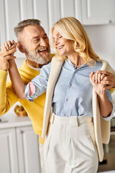 A mature, loving couple dances in a cozy kitchen, enjoying each others company and the music in their comfortable homewear.