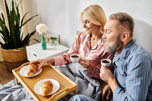 A mature couple in cozy homewear sitting on a couch, sipping coffee and indulging in pastries together in a warm and inviting atmosphere.