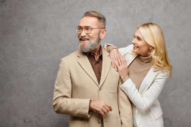 Olgun, zarif bir erkek ve kadın güler yüzlü bir şekilde yan yana durur. Gri bir zemine karşı sevgi ve entelektüellik yayar..