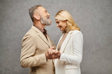 Mature, elegant couple in debonair attire embrace, holding hands lovingly against a gray backdrop. clipart
