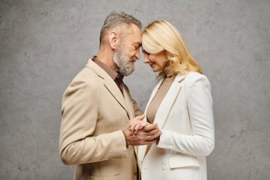 Olgun erkek ve kadın, zarif giyinmiş, gri bir arkaplanda sevgi dolu bir pozda yan yana dururlar..