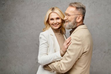 Zarif giyinmiş olgun bir erkek ve kadın gri bir zemine nazikçe sarılır..