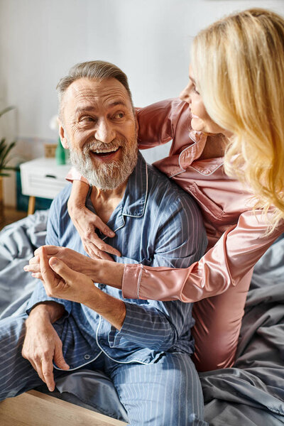 A woman sits on a bed next to a man in cozy homewear, sharing a moment of intimacy and connection.