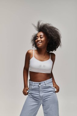 Kıvırcık saçlı, güzel bir Afro-Amerikan kadın beyaz bluz ve kot pantolon içinde rahatça poz veriyor..