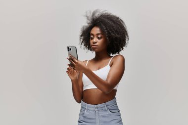 Çekici, kıvırcık saçlı Afrikalı Amerikalı bir kadın stüdyo ortamında beyaz bir bluz giyerken cep telefonu tutuyor..