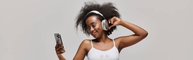 A young African American woman with curly hair holds a cell phone and wears headphones, absorbed in the digital world. clipart