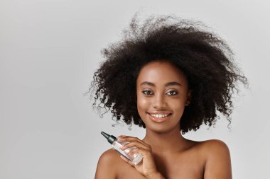 Afro saçlı, çarpıcı bir Afrikalı Amerikalı kadın elinde şişeyle doğal güzelliğini kucaklıyor..