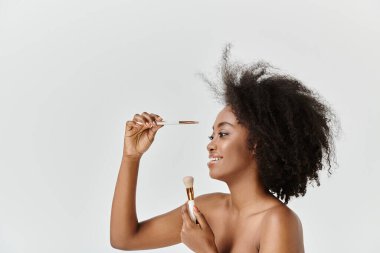 A young African American woman with curly hair applying a brush to her face as part of a skin care routine. clipart