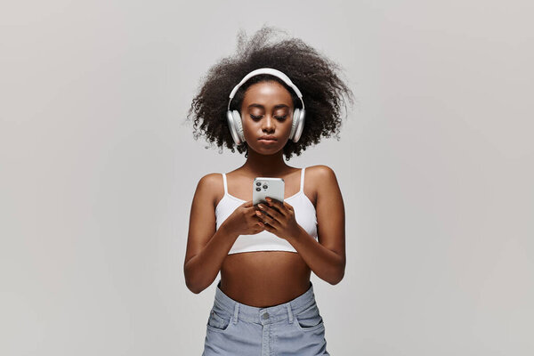 A young African American woman with curly hair, wearing headphones, gazes at her cell phone screen attentively.