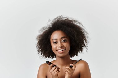 Gülümseyen Afrikalı Amerikalı bir kadın, stüdyo ortamında maskara sürüyor, neşe ve yaratıcılık saçıyor..