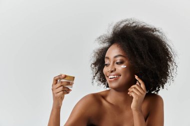 Kıvırcık saçlı genç bir Afrikalı Amerikalı kadın elinde krema kavanozuyla gülümsüyor, neşe ve memnuniyet saçıyor..