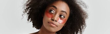 Çarpıcı kıvırcık saçlı Afro-Amerikan bir kadın çarpıcı bir kırmızı göz bandı takarken cilt bakımı konseptini somutlaştırıyor..