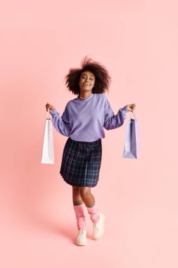 Kıvırcık saçlı, mor kazaklı ve ekose etekli genç bir Afro-Amerikalı kadın, şık bir stüdyo fotoğraf çekiminde alışveriş torbalarını tutuyor..