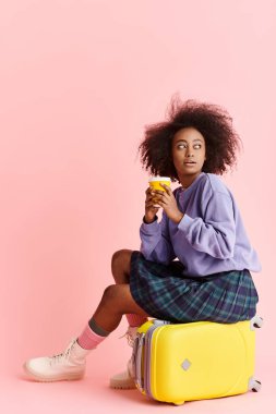 Kıvırcık saçlı genç Afrikalı Amerikalı bir kadın stüdyoda canlı sarı bir valizin üzerinde zarif bir şekilde oturuyor..