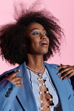 Şık, kıvırcık saçlı, mavi takım elbiseli Afro-Amerikan bir kadın stüdyo ortamında güven içinde poz veriyor..