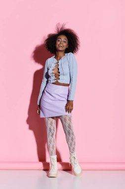 A stylish young African American woman with curly hair strikes a pose in front of a vibrant pink wall. clipart