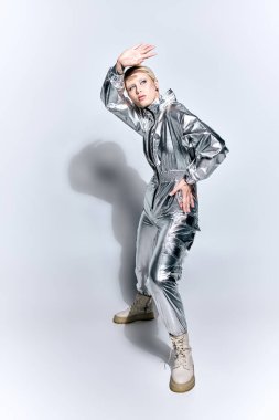 Gümüş rengi fütüristik elbiseli çekici bir kadın gri arka planda poz veriyor ve başka tarafa bakıyor.