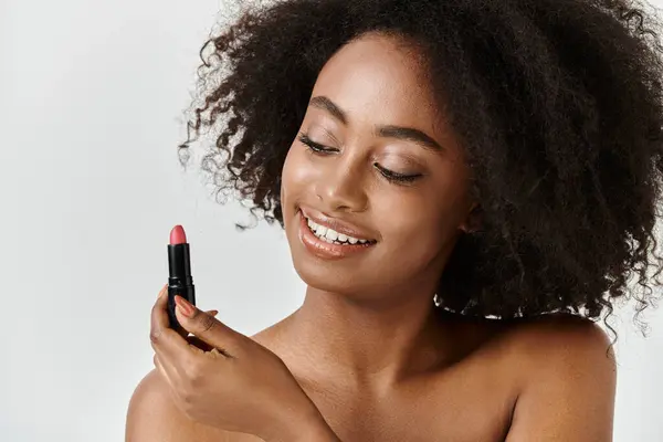 stock image A beautiful young African American woman with curly hair holds a lipstick in her hand, embodying a skin care concept.