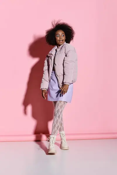 stock image A stylish young African American woman with curly hair stands confidently in front of a vibrant pink wall.